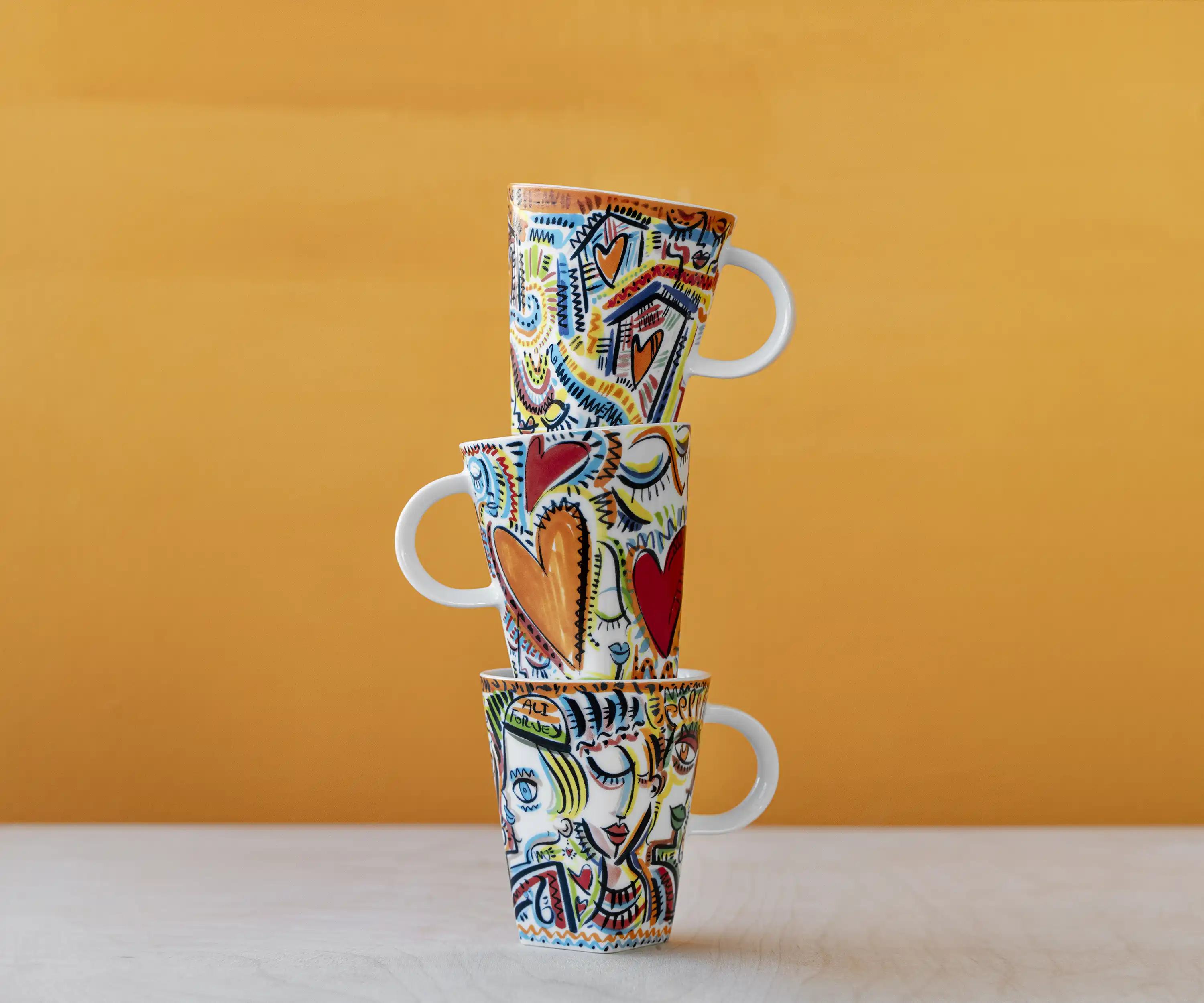 Three stacked Nespresso cups painted with colorful hearts and faces on a mustard backdrop, Things We Hold artist cups