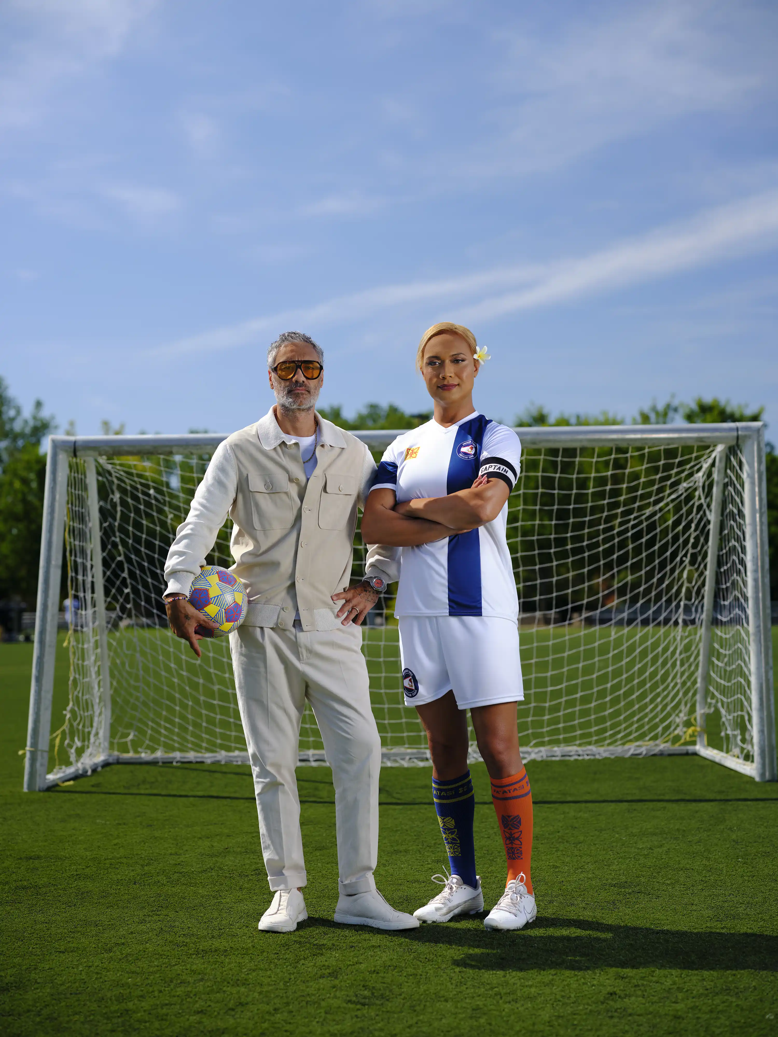Taika Waititi holds a patterned soccer ball beside Jaiyah Saelua in a white-and-blue kit, posed at a goal for Next Goal Wins