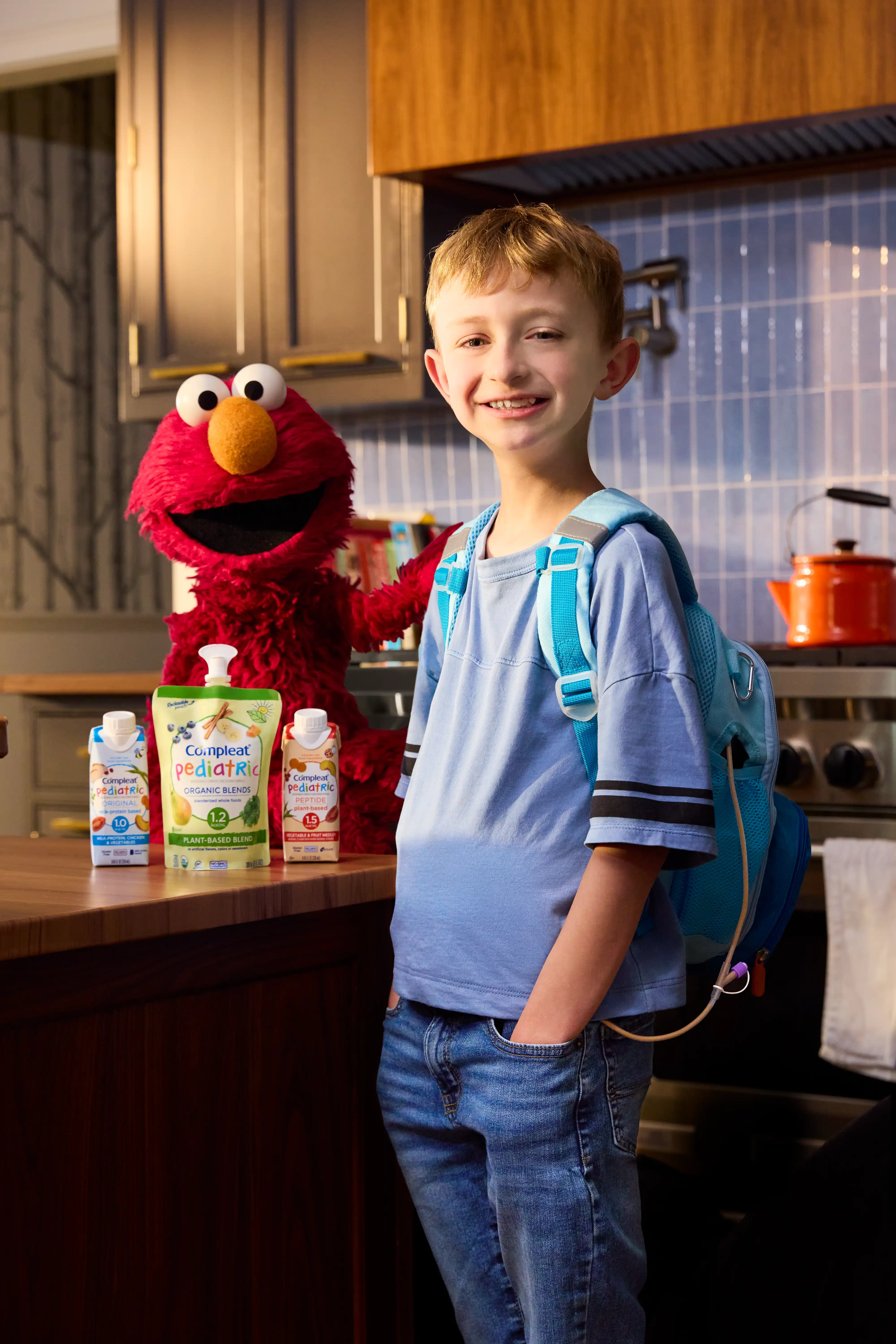 A smiling boy with a feeding backpack stands in a kitchen with Elmo and Compleat Pediatric cartons