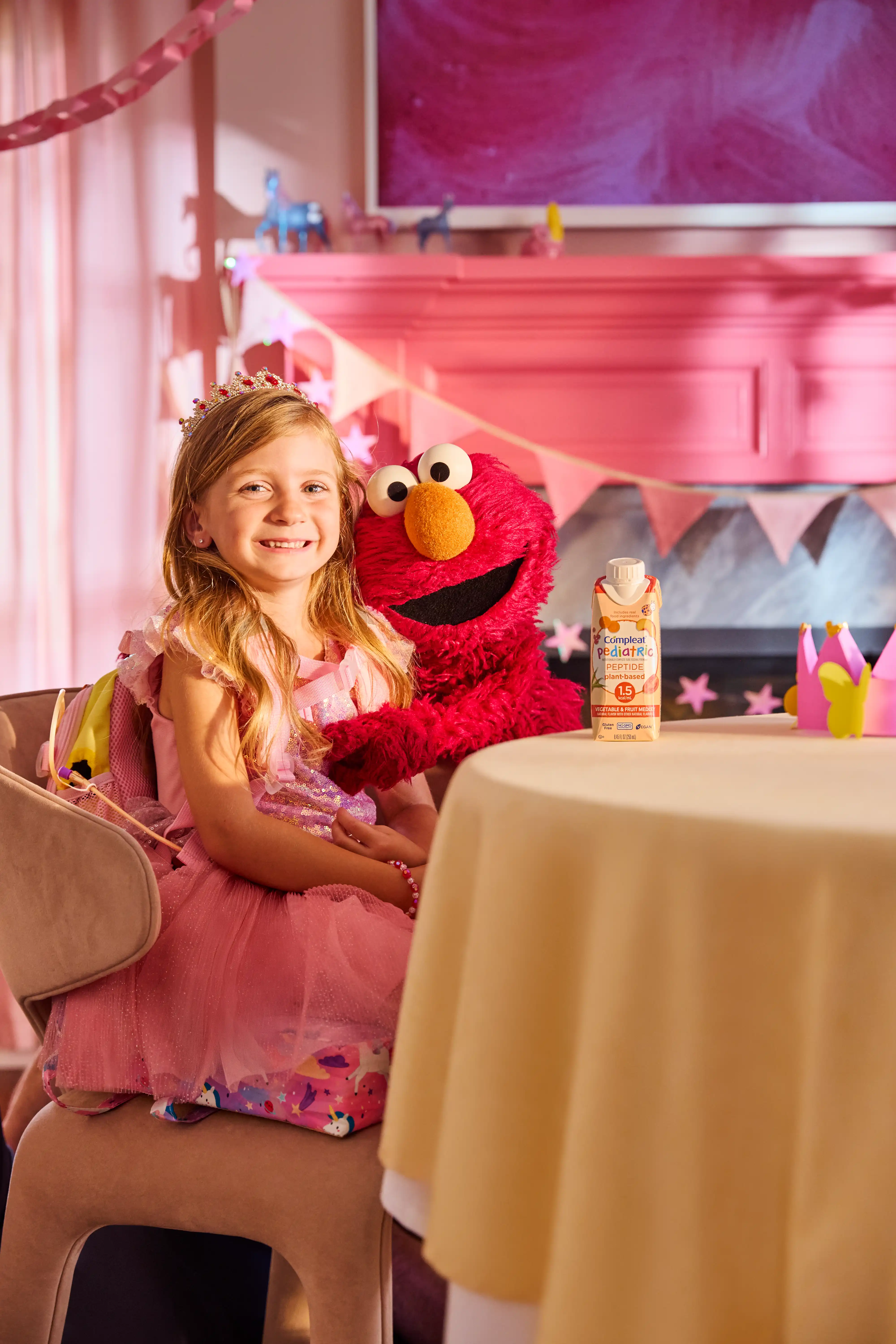 A girl in a pink princess dress sits with Elmo and a Compleat Pediatric carton at a party table
