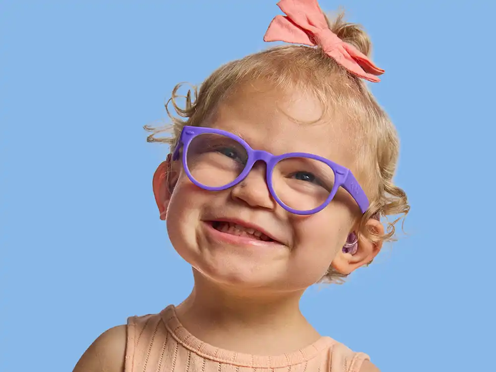 A toddler in purple glasses and a pink bow smiles against blue, from Compleat x Elmo's What Is Better Than How