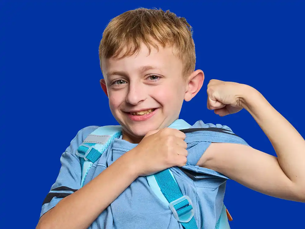 A boy in a blue feeding backpack flexes his arm against a blue backdrop, from Compleat x Elmo's What Is Better Than How