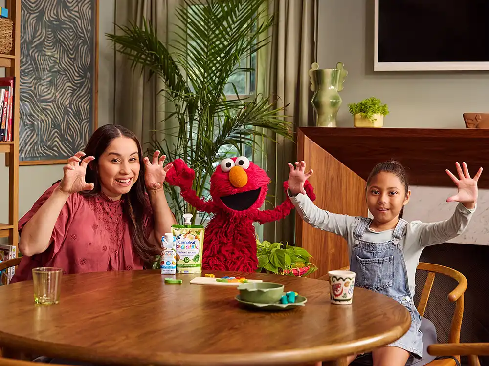 A mother, Elmo, and a girl in overalls raise their hands at a table with Compleat Pediatric, from What Is Better Than How