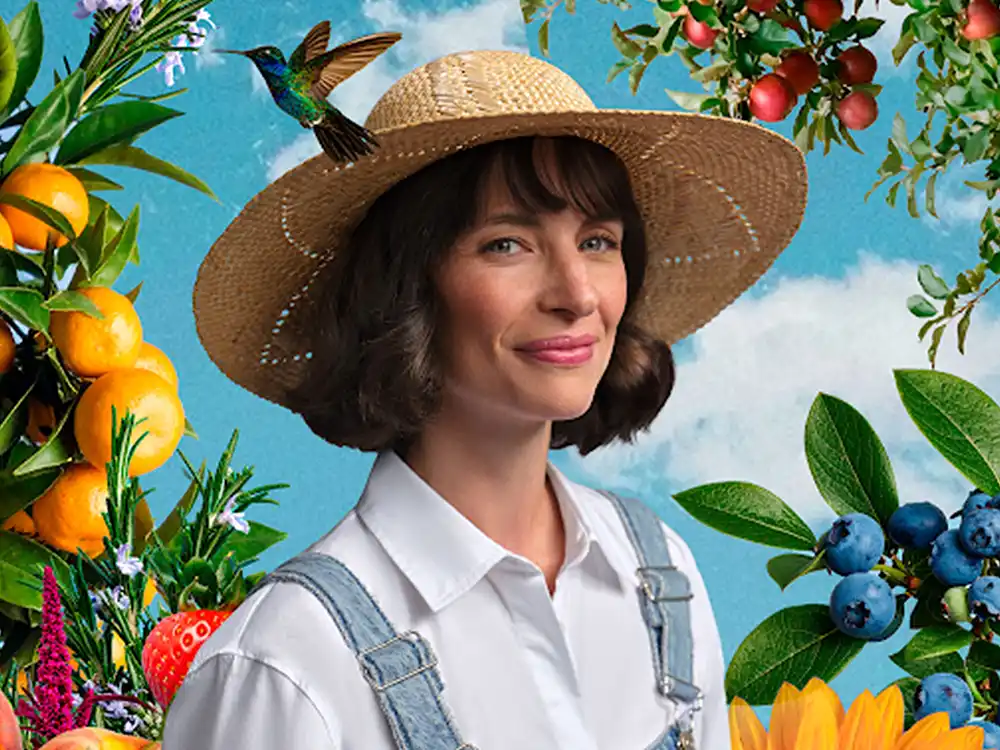 A woman in a straw hat and overalls smiles among citrus and blueberries, a hummingbird on her brim, from Garden of Life's Formulas for Feeling Alive