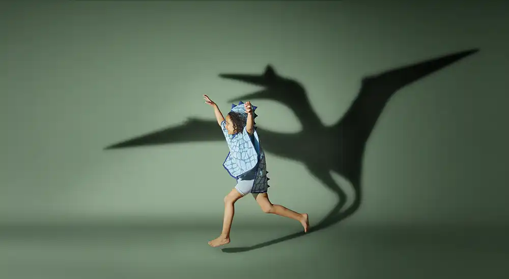 Child in a blue dinosaur hooded towel runs across a green studio set, casting a pterodactyl shadow