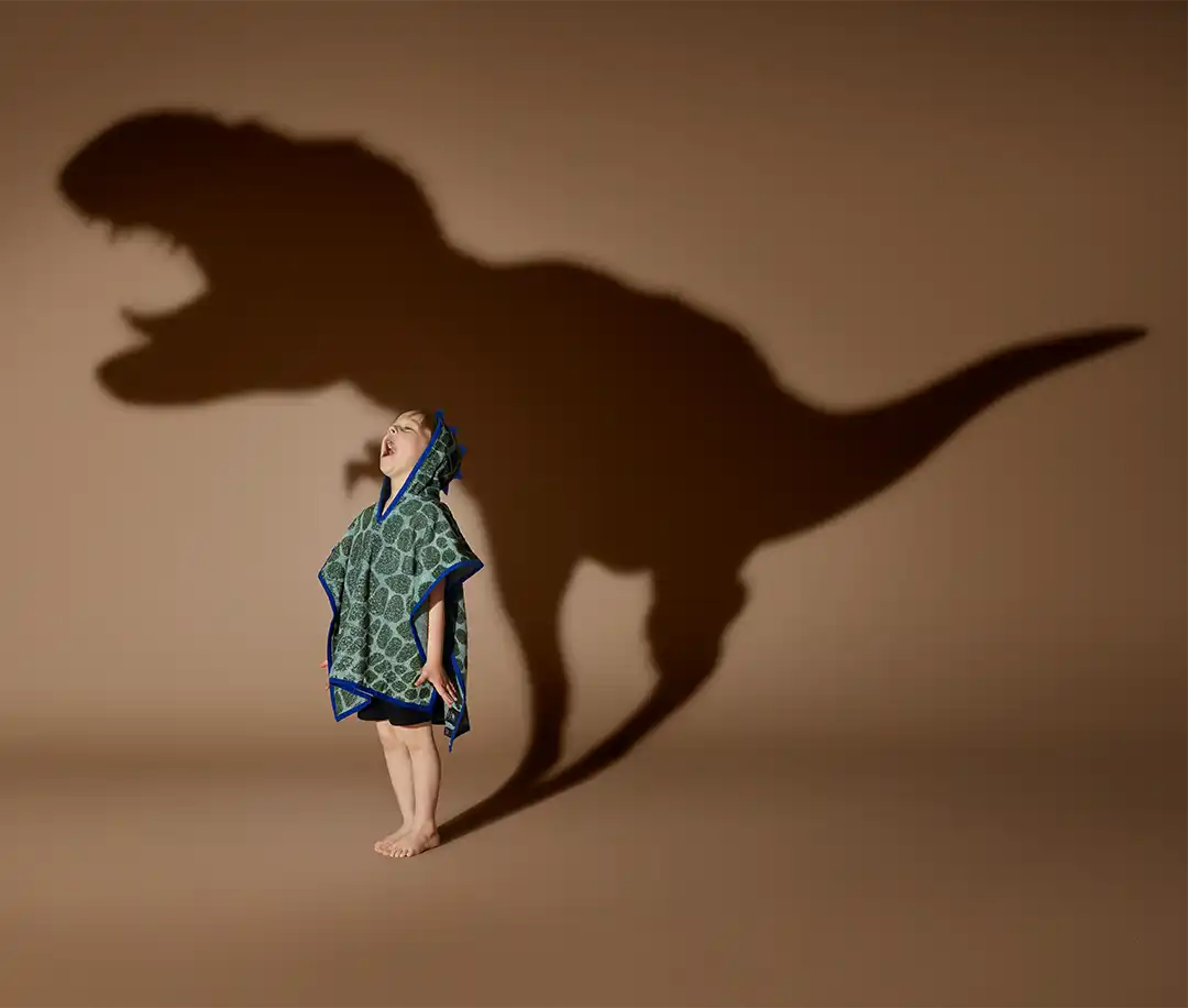 Small child in a green dinosaur hooded towel looks up at a huge T. rex shadow on a brown studio backdrop