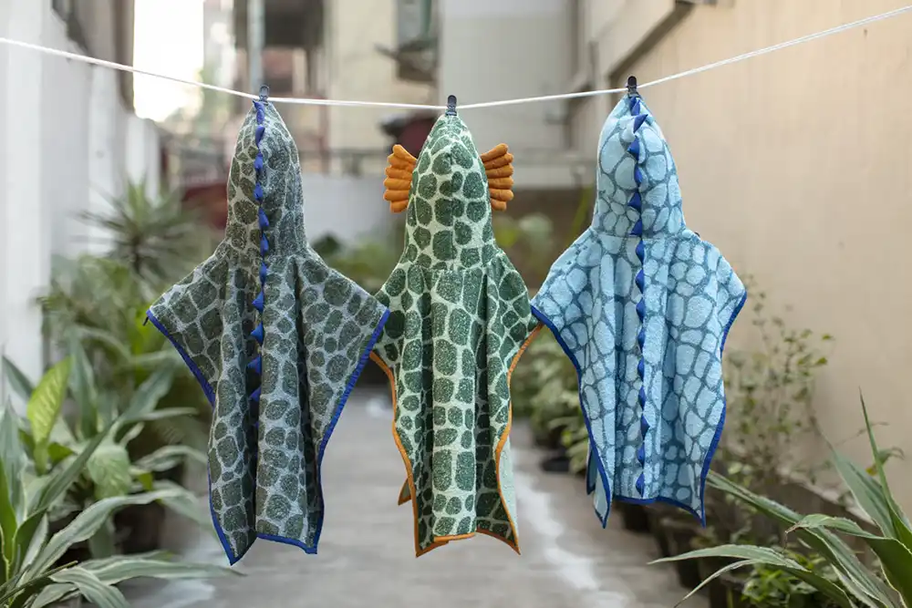 Three dinosaur-print hooded towels hang on a clothesline in an alley—green, orange-eared, and blue