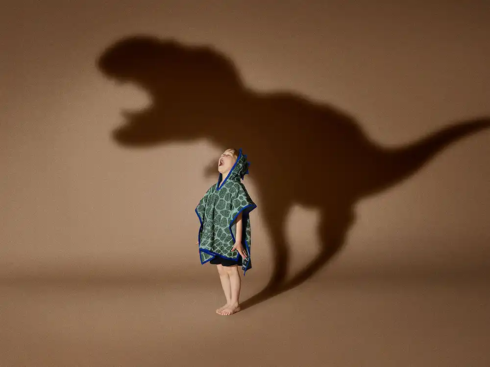Child in a green dinosaur hooded towel looks up at a T. rex shadow, from Jurassic World's Preserving Legacies of the Past collection