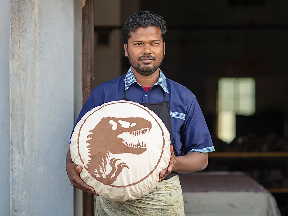 Artisan in a workshop holds a round cream pillow with a brown T. rex logo, from Jurassic World's Preserving Legacies of the Past collection