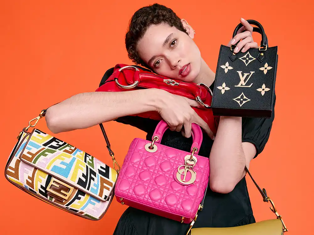 Person cradles Fendi, Dior, and Louis Vuitton bags against orange, from Vestiaire Collective's Parlez-vous?