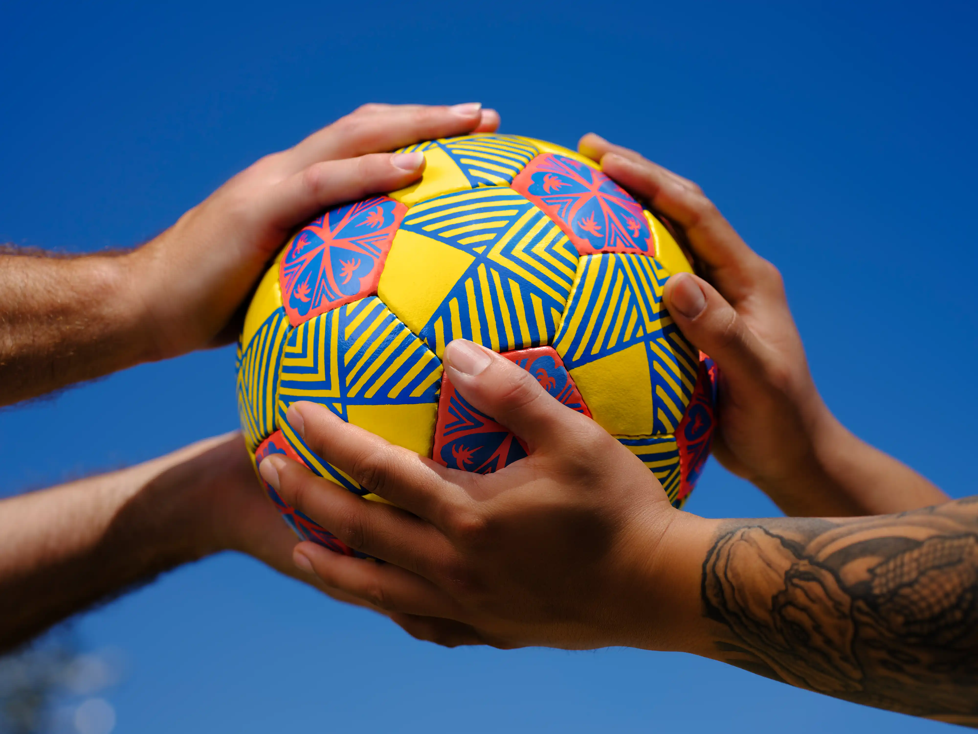 Hands holding the Fa'atasi patterned soccer ball against a blue sky